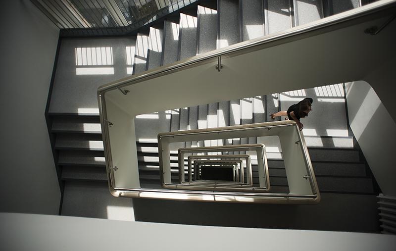 Widok z góry na klatkę schodową. Schody ułożone prostokątnie. Kobieta w czarnej sukience wchodząca po schodach.
