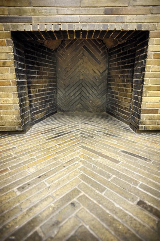 Wnętrze kominka - otwór w ścianie wyłożony brązowymi wąskimi płytkami ułożonymi poziomo. Na podłodze i centralnej ścianie płytki ułożone w jodełkę.